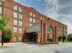 A four-story brick hotel building with large arched windows and a covered entrance. The sky is partly cloudy, and there is a small parking area in front.