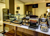 Buffet breakfast area with pastries, bread, a toaster, and condiments on a counter. Signs label items such as toast and syrup.