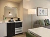 Hotel room kitchenette with a mini-fridge, microwave, and coffee maker next to a small table. A couch with patterned cushions and a tall floor lamp are visible. Art hangs on the wall.