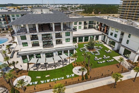 Aerial view of a large, modern hotel with a pool, outdoor seating areas, and landscaped gardens, set against a backdrop of trees and neighboring buildings.