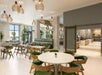 Modern cafe interior with round tables and green chairs, natural light from large windows, decorative hanging lights, and wall mirrors.