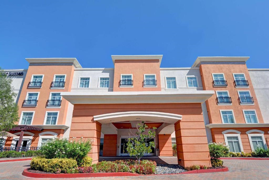 A three-story hotel with a red and beige facade, black railings on balconies, and a covered entrance, surrounded by landscaped greenery under a clear blue sky.