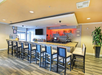 Hotel bar area with a granite countertop, high chairs, a range of bottled drinks on shelves, a TV screen, and a decorative plant.