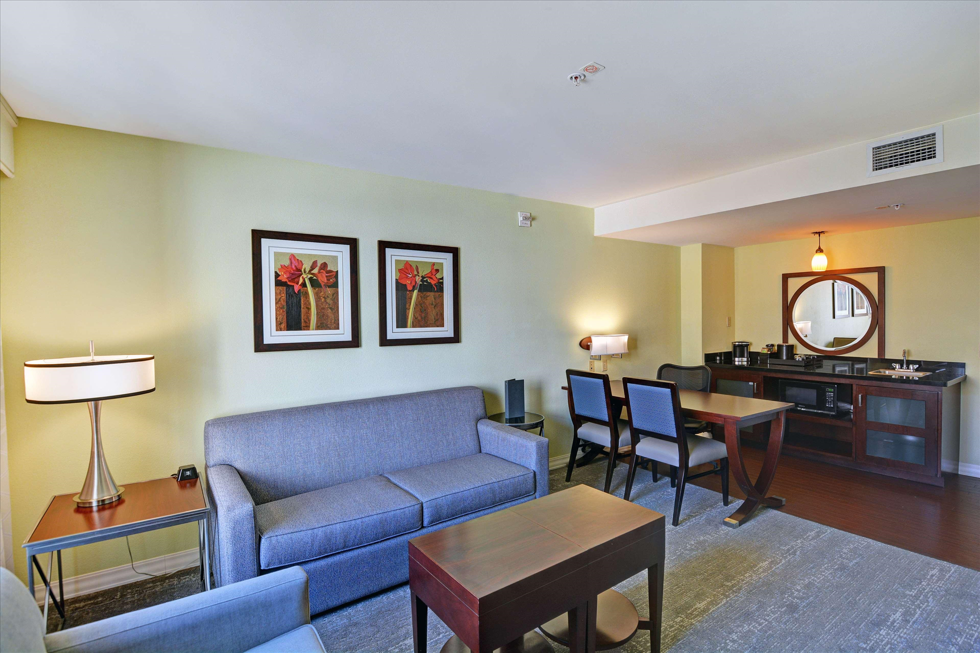 A modern hotel room with a seating area, featuring a gray sofa, two framed floral artworks, a wooden table, and a minibar with chairs. Walls are cream-colored, and lighting is soft.