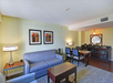 A modern hotel room with a seating area, featuring a gray sofa, two framed floral artworks, a wooden table, and a minibar with chairs. Walls are cream-colored, and lighting is soft.