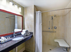 Accessible bathroom with a large mirror, sink, toiletries, and towels on the left. Shower area with a curtain, handrails, and foldable bench on the right.