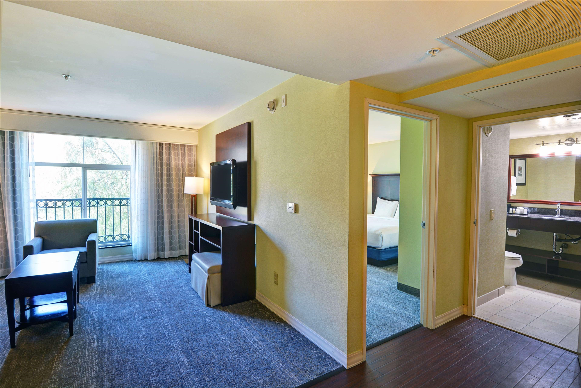 Spacious hotel room with a seating area, TV, and desk. Bedroom and bathroom with modern amenities are visible. Large window with curtain and balcony railings in the background.