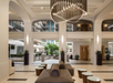 Spacious hotel lobby with modern chandelier, beige seating, and glass coffee tables. Arched balconies overlook the area, and potted plants are arranged throughout.