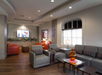 Modern lounge area with gray sofas, orange armchairs, a large window, and a wall-mounted TV. Wooden floor and a table with an orange vase complete the decor.