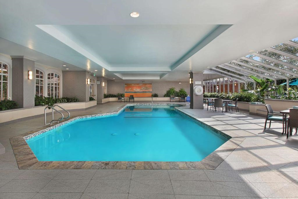 Indoor swimming pool with tiled floor, surrounded by windows and greenery. Tables and chairs are set around the pool area.