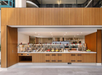 Modern cafeteria with a self-serve food area featuring a wooden counter, metal dispensers, and various dishes. Bright overhead lighting and clean design.