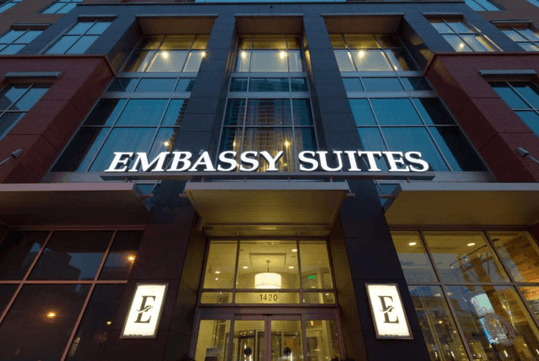 The bright white sign for the Embassy Suites above the glass front entrance in Denver, Colarado.