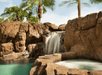 A rocky waterfall flows into a turquoise pool, surrounded by palm trees under a clear sky.