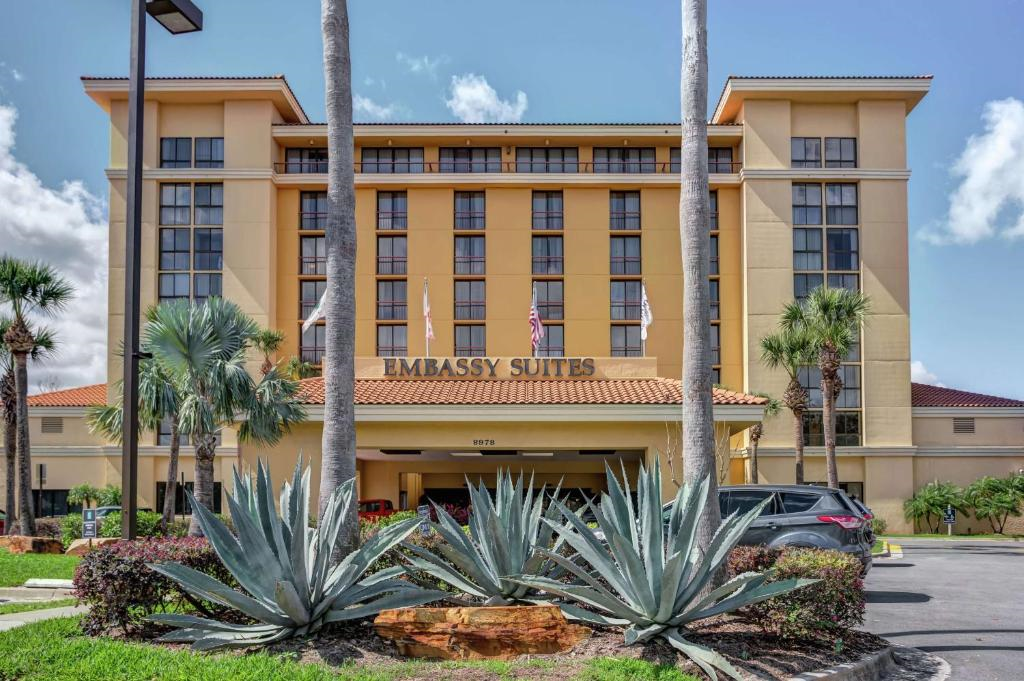 Front view of Embassy Suites hotel with tall palm trees, multiple flagpoles, and a neatly landscaped entrance featuring large agave plants.