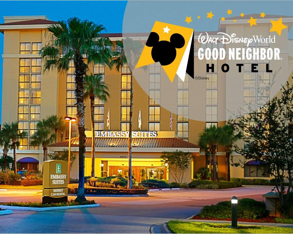 Exterior view of Embassy Suites hotel at dusk, featuring palm trees and lights, with a Walt Disney World Good Neighbor Hotel logo in the corner.