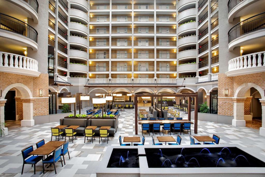 Spacious hotel atrium with multiple floors, featuring dining tables, chairs, decorative fountains, and a checkered floor. Balconies line each level.
