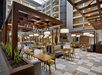 Modern hotel indoor dining area with wooden furniture, hanging lights, and green plants. Tables and chairs are arranged neatly under a wooden canopy, with balconies visible in the background.