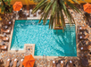 Aerial view of a rectangular swimming pool surrounded by lounge chairs and orange umbrellas on a tiled patio, with a palm tree partly covering the pool.