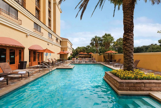 Outdoor hotel swimming pool with lounge chairs, umbrellas, and palm trees in a landscaped area. Building with rows of windows in the background on a sunny day.
