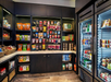 A small convenience store featuring shelves with packaged snacks, drinks, and a refrigerated section on the right displaying cold beverages and snacks.