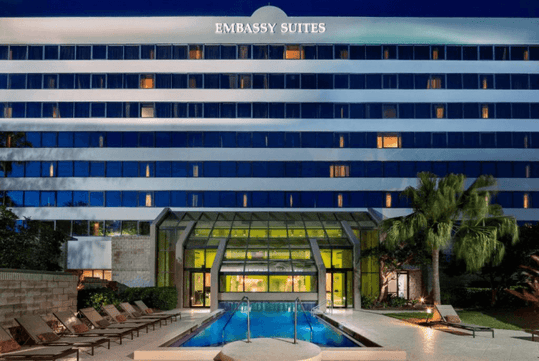 A small bright blue outdoor swimming pool with lounge chairs on the left and a palm tree on the right with the Embassy Suites behind it.
