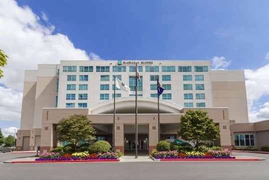 The front exterior of the Embassy Suites by Hilton Portland Airport  with a tree, bushes, and other landscaping on both sides of the walkway on a sunny day.