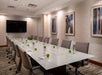 A meeting room with a long white glass topped table set with pen, paper, water, and green apples with a TV at the far end of the room and paintings on the right wall.