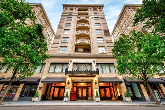 The front exterior of the Embassy Suites by Hilton Portland Downtown, a tall beige brick building with glass doors on the bottom and trees on the sides.