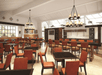 Spacious restaurant interior with orange chairs, dark tables, and chandeliers. Large windows and a high ceiling allow natural light in.