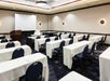 A conference room with rows of white cloth covered tables with black chairs set up in front of a large projector and a podium.