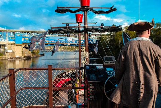 A person dressed as a pirate steers a ship decorated with a skeleton and flags, traveling under a blue bridge on a sunny day.
