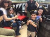 Adults and children wearing pirate hats sit and stand on a boat deck, with one child held by an adult and two children posing near a wooden barrel.