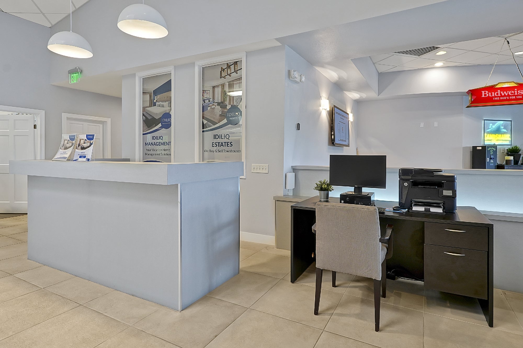 A modern office reception area with a front desk, a workstation with a computer and printer, promotional posters, and pendant lights.