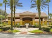 A building with a tiled roof and yellow columns labeled "Encantada," surrounded by palm trees, shrubs, and flowers under a partly cloudy sky.