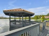 A wooden gazebo with a red roof extends over a lake, connected by a walkway, with buildings and trees in the background under a blue sky.