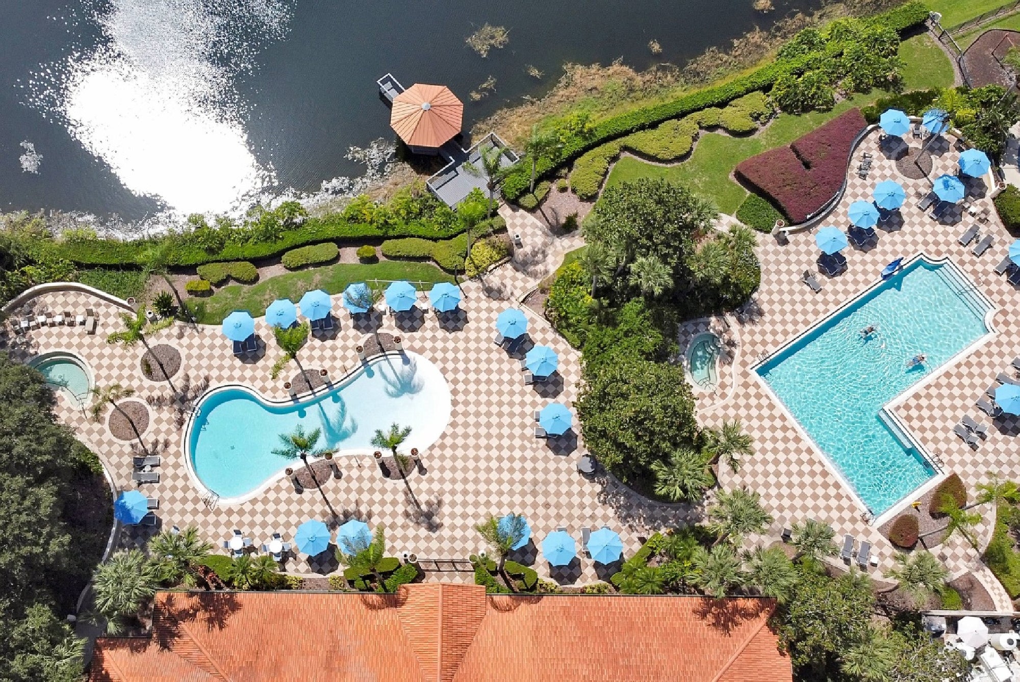 Aerial view of a pool area with two pools, blue umbrellas, lounge chairs, landscaped greenery, and a dock by a body of water.