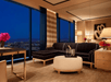 Luxurious living room with a black sectional sofa, round ottoman, and large windows overlooking a cityscape at night. A vase with flowers and a lamp are on a nearby table.