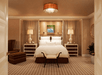 Luxurious hotel bedroom with a central white bed, flanked by two nightstands and lamps. A bench sits at the foot of the bed, and a patterned carpet covers the floor.