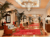 Elegant hotel lobby with plush seating, red carpet, palm plants, and ornate chandeliers. Walls feature paintings and decorative elements, creating a luxurious atmosphere.