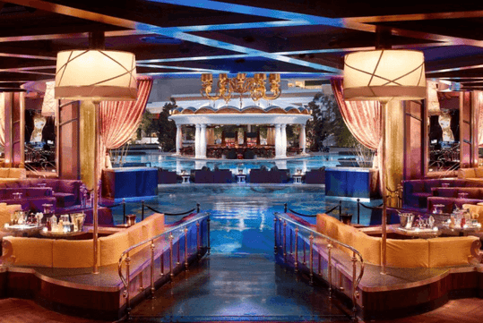 Luxurious poolside lounge with plush seating, two large lamps, draped curtains, and a pool bordered by elegant structures in the background.