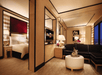 Luxurious hotel suite featuring a bedroom with a king-size bed, a living area with a black sofa, a TV, and a coffee table with a champagne bottle and fruit.