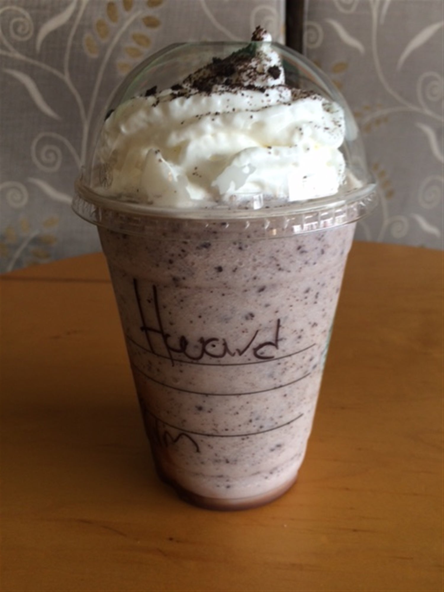 A plastic cup of cookies and cream milkshake topped with whipped cream and crushed cookies sits on a wooden table.