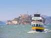 A ferry labeled "Blue & Gold Fleet" approaches, with passengers on deck. Alcatraz Island is visible in the background under a clear sky.