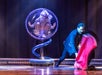 A woman in a silver costume sitting in a clear bubble on a pedestal with a man in all black having just pulled a red sheet off her.
