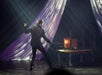 A man in an all black suit with one of his hands up and the other holding a stick and lighting a box on fire on stage.