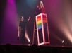 A man in all black holding a book in one hand and point to a woman standing in a rainbow box above him on stage.