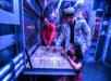 Three people in a submarine-themed room examine a large map on a table; one person wears a captain’s uniform and points to the map.