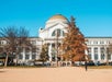 The image shows the Smithsonian National Museum of Natural History, a neoclassical building with a large dome, on a clear day with people walking outside.