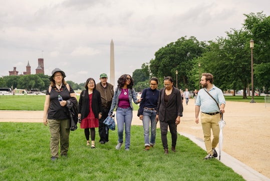 A group of six adults walk and talk together on a grassy area near the Washington Monument on a cloudy day.