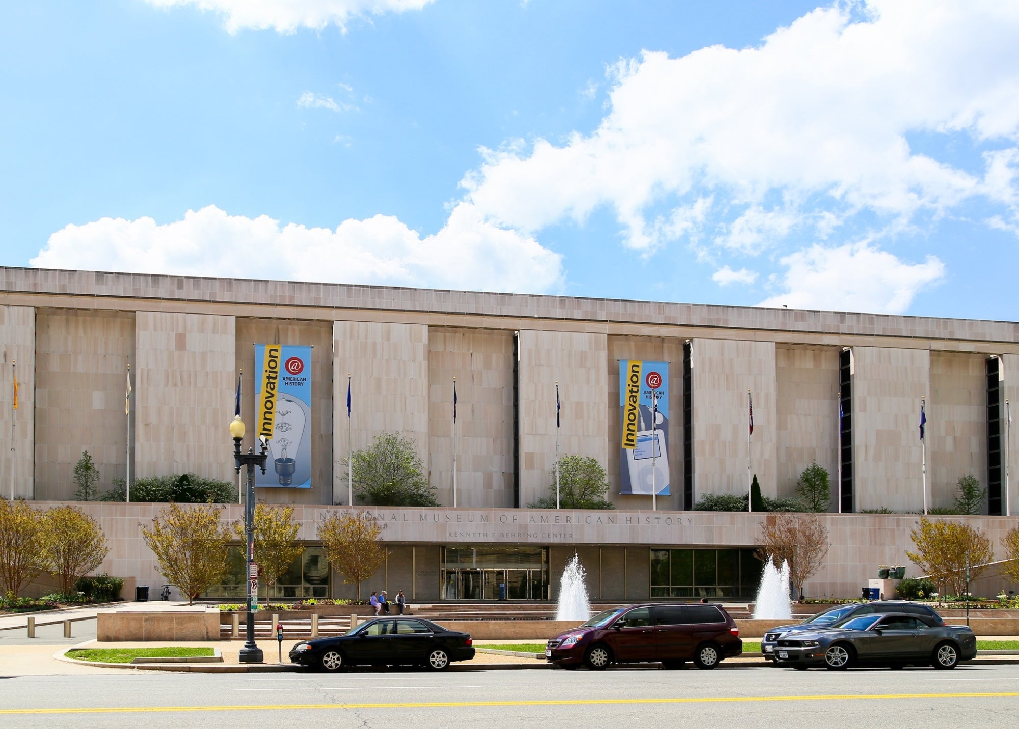 Exterior view of the National Museum of American History with banners reading "Innovation," fountains, parked cars, and trees in front of the building.